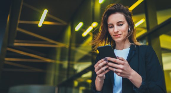 Lifestyle portrait of happy business woman walking in evening city and using mobile gadget, beautiful young girl banker dressed in business style listening to music wireless earphones and typing text message on smartphone device, flare light